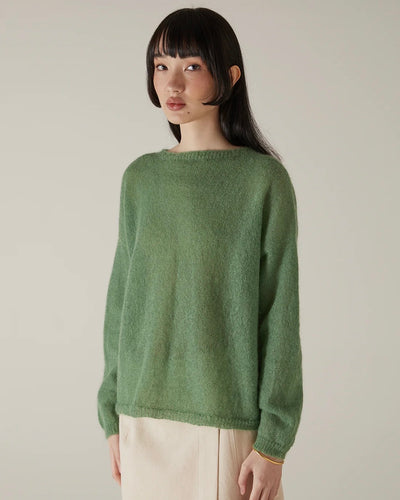 A person with straight dark hair wears the Feather Knit – Clover, a relaxed fit green sweater, paired with beige pants, standing against a plain light background.