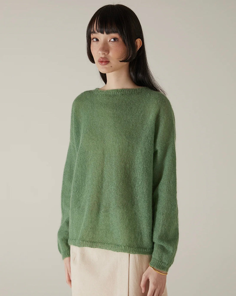 A person with straight dark hair wears the Feather Knit – Clover, a relaxed fit green sweater, paired with beige pants, standing against a plain light background.