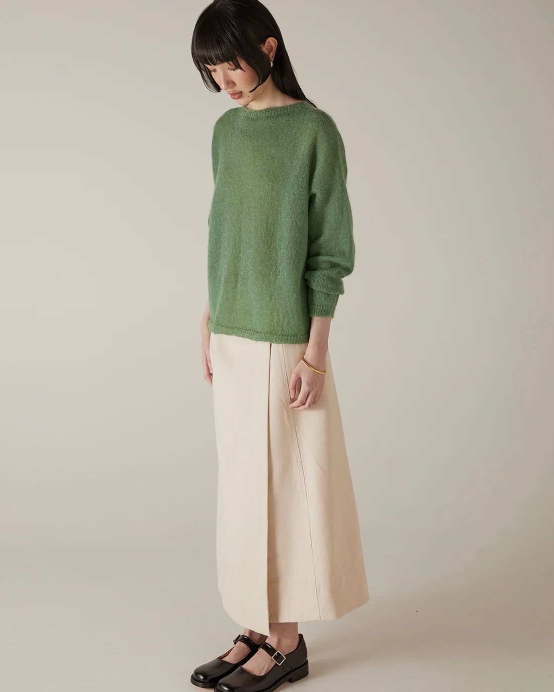 A woman stands against a plain background, wearing the Feather Knit – Clover sweater, paired with a long beige skirt and black Mary Jane shoes. She looks slightly downward with her hands at her sides.