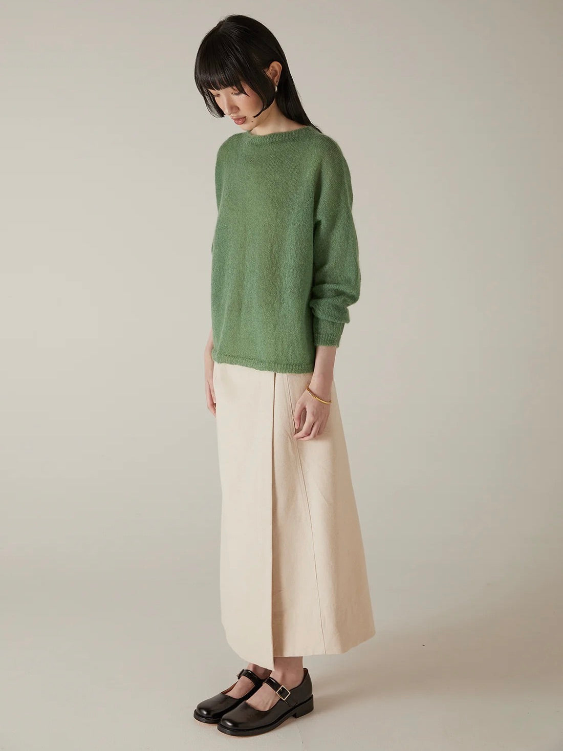 A woman stands against a plain background, wearing the Feather Knit – Clover sweater, paired with a long beige skirt and black Mary Jane shoes. She looks slightly downward with her hands at her sides.