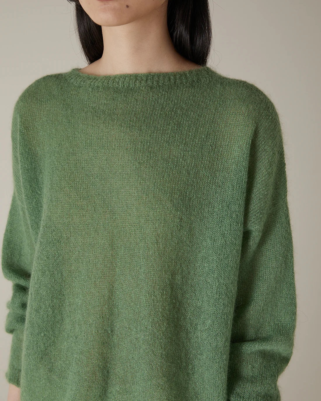 A person with shoulder-length dark hair models the Feather Knit – Clover, a relaxed fit, long-sleeved green sweater, shown from shoulder to mid-thigh against a plain background.