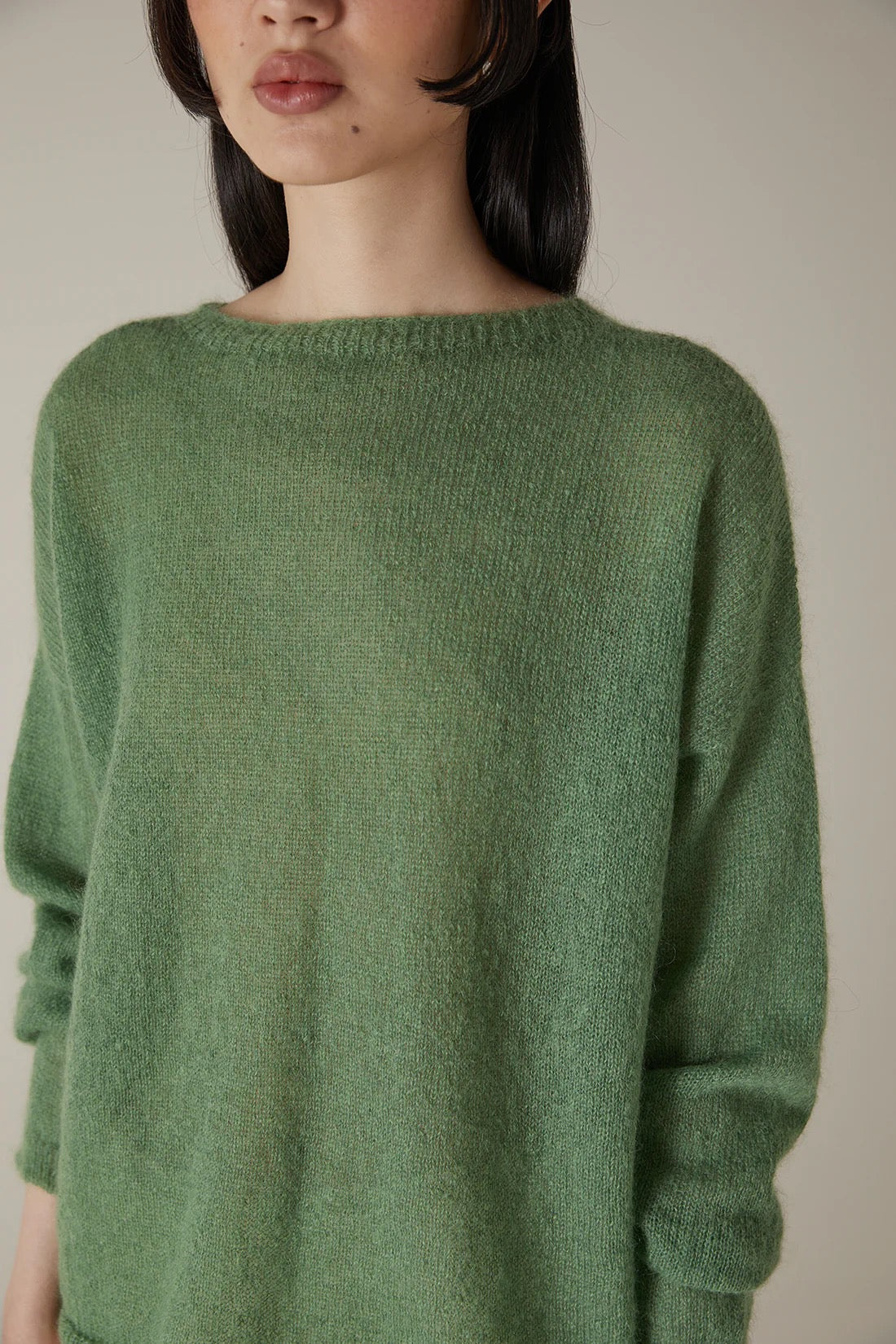 A person with shoulder-length dark hair models the Feather Knit – Clover, a relaxed fit, long-sleeved green sweater, shown from shoulder to mid-thigh against a plain background.