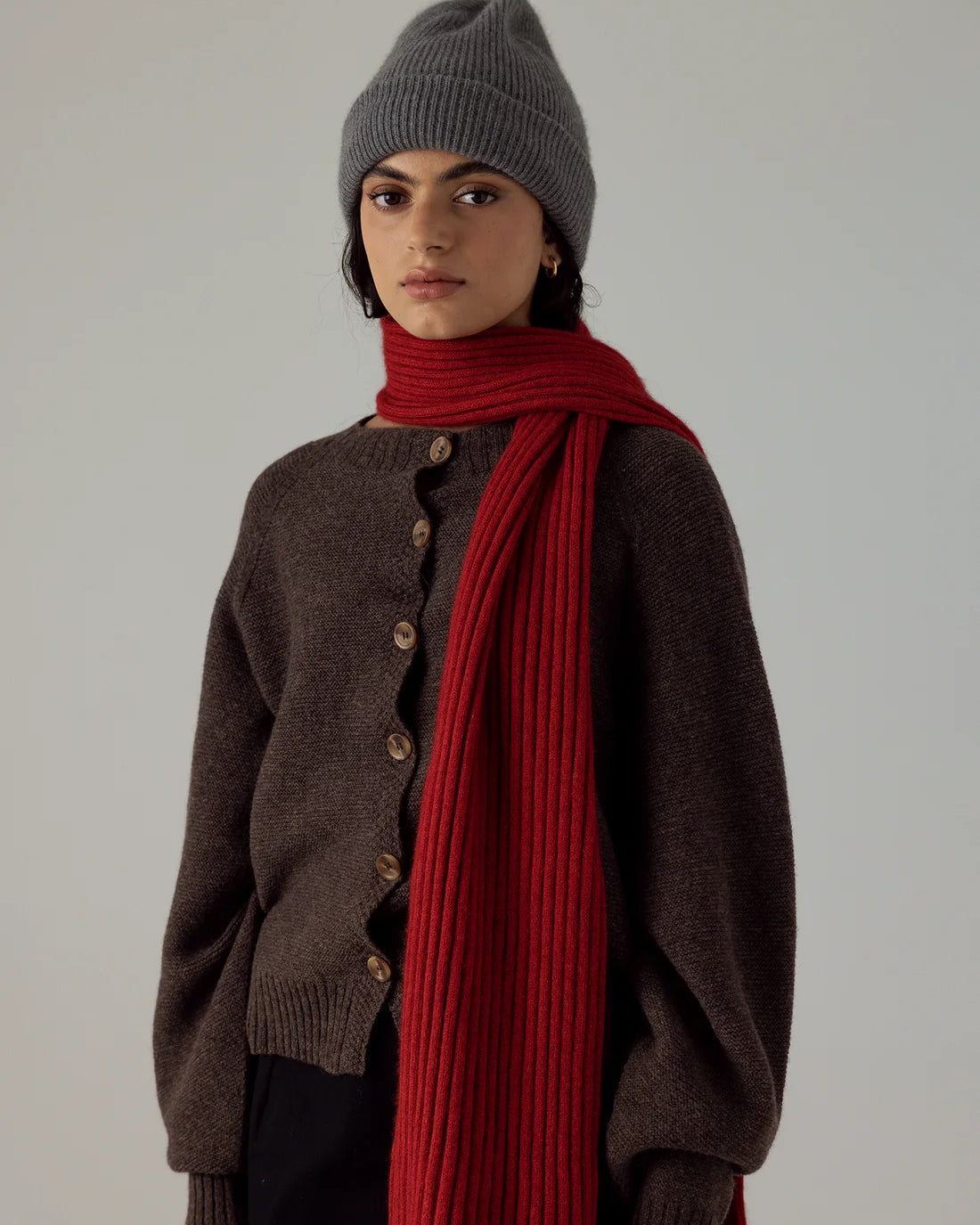 A person wears the Hinoki Beanie in Granite, a brown button-up sweater, and a long red scarf, standing against a plain light background.