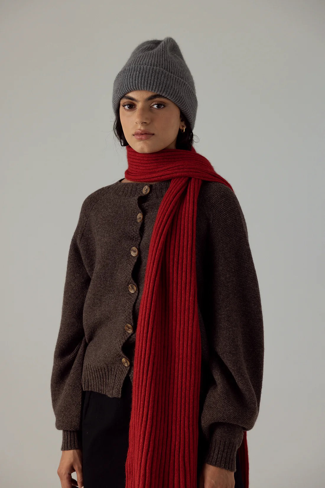 A person wears the Hinoki Beanie in Granite, a brown button-up sweater, and a long red scarf, standing against a plain light background.