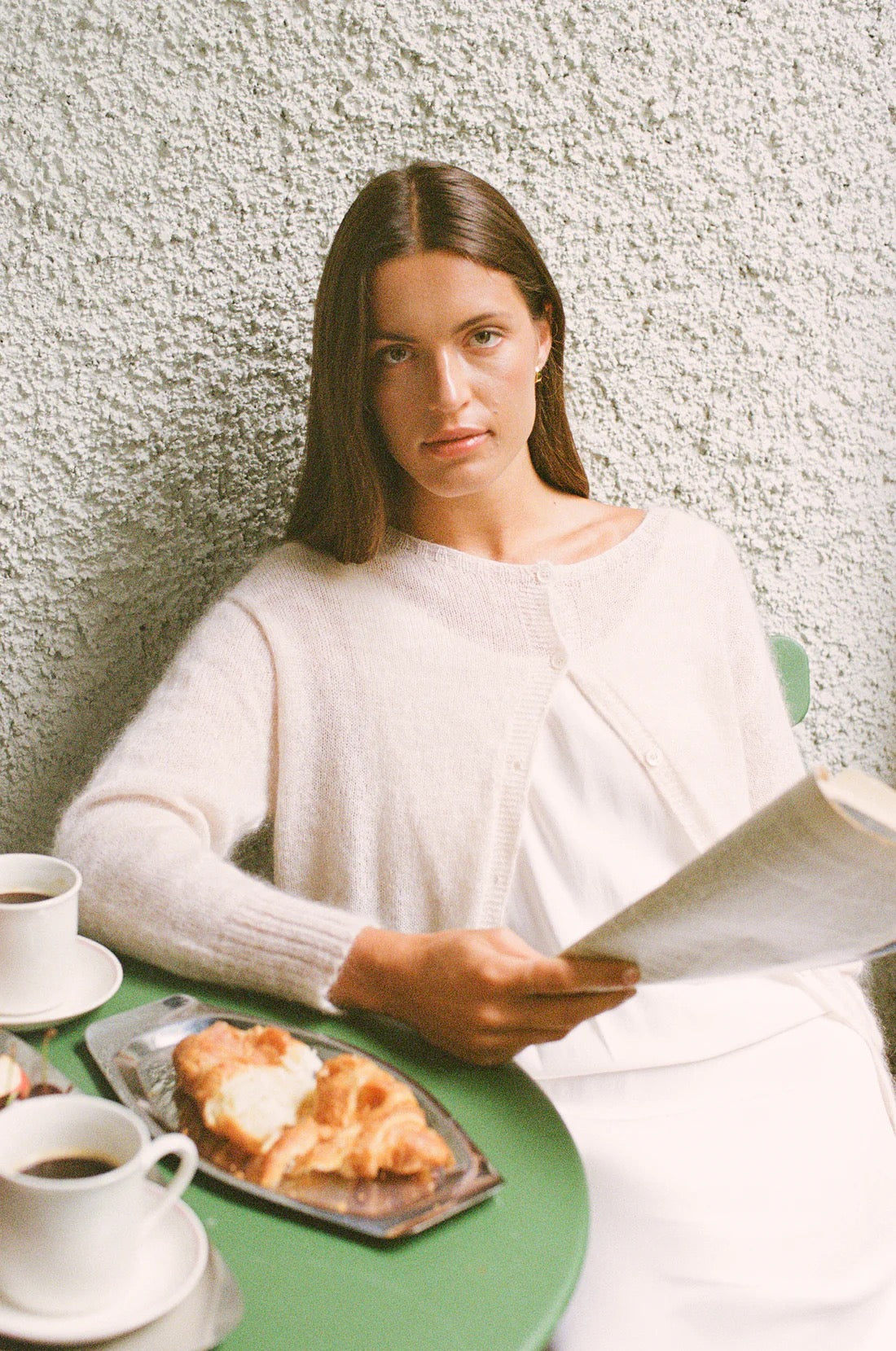 A woman in a relaxed fit Fleur Cardigan – Creme sits at a green table with coffee, pastries, and an open book against a textured white wall.