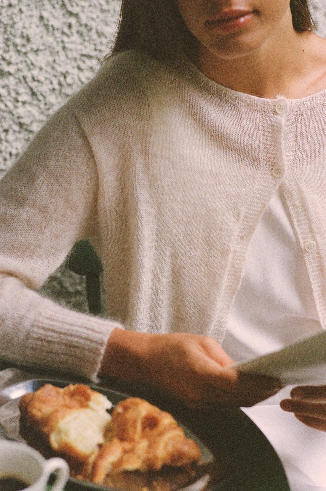 Wearing the Fleur Cardigan – Creme, a person with a relaxed fit sits at a table, holding paper, with croissants and coffee placed before them.
