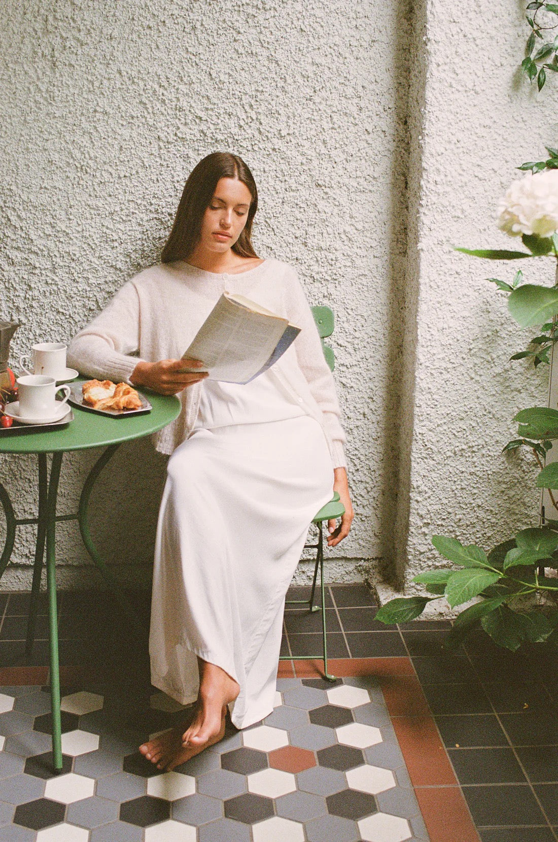 A petite woman sits barefoot at a small green table, reading with breakfast nearby, wearing the Fleur Cardigan – Creme over a long white skirt in a bright, textured indoor space.