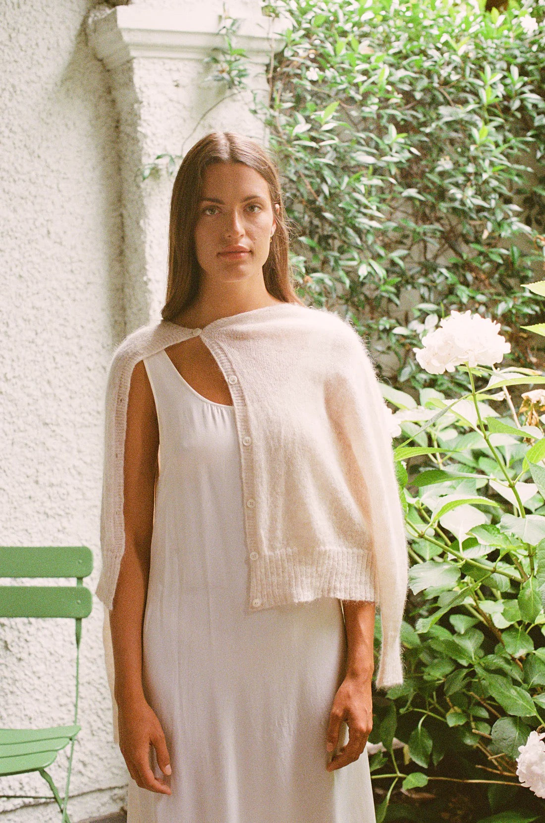 A petite woman wearing the Fleur Cardigan in Creme over a white dress stands in a garden beside green foliage and a white flower.