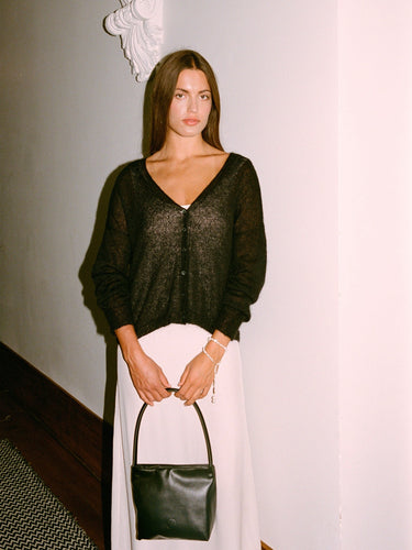A petite woman stands indoors against a white wall, wearing the Mist Cardigan in black, a white skirt, and holding a black handbag.