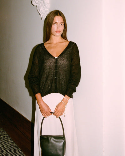 A petite woman stands indoors against a white wall, wearing the Mist Cardigan in black, a white skirt, and holding a black handbag.