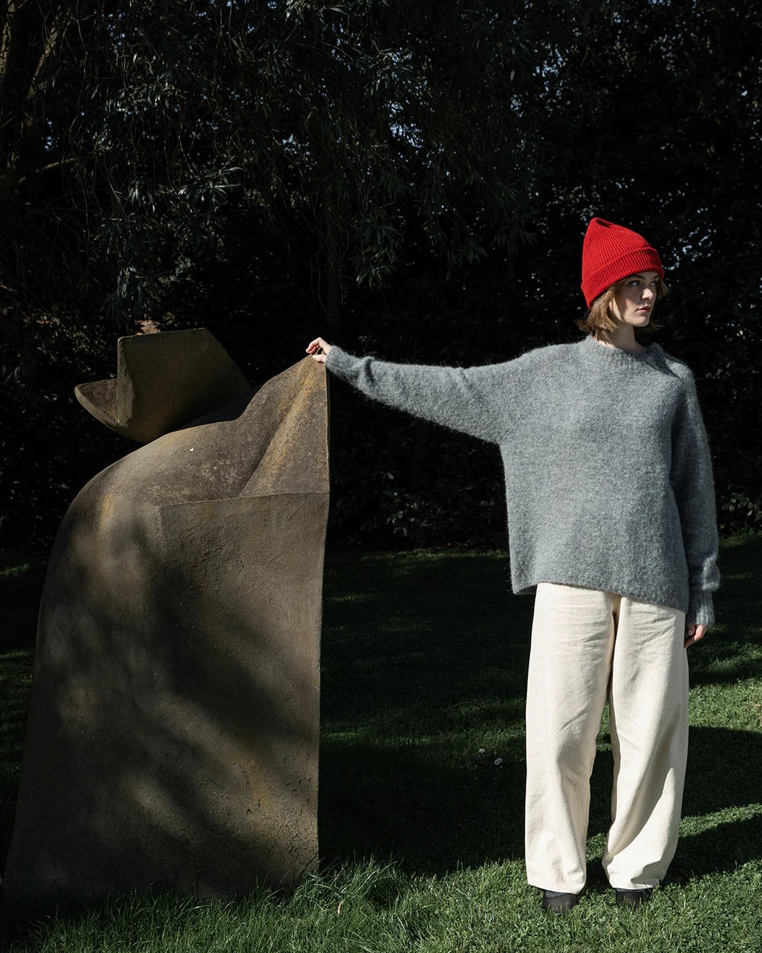 A person in a gray sweater, white pants, and the Hinoki Beanie – Poppy stands on grass, touching a large abstract stone sculpture outdoors.