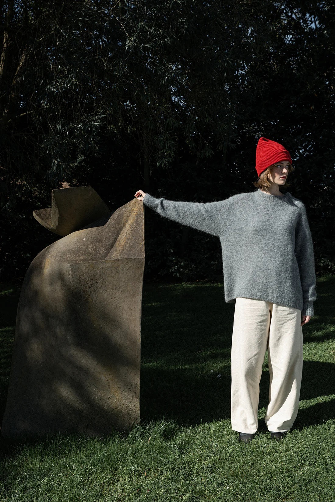 A person in a gray sweater, white pants, and the Hinoki Beanie – Poppy stands on grass, touching a large abstract stone sculpture outdoors.