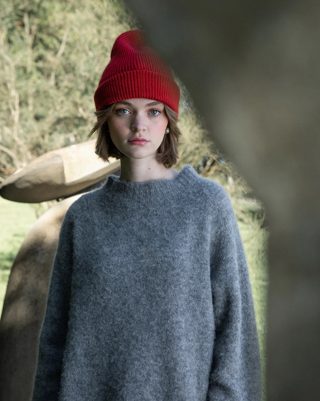 A person wearing the Hinoki Beanie – Poppy and a loose gray sweater stands outdoors near abstract stone sculptures, with trees in the background.