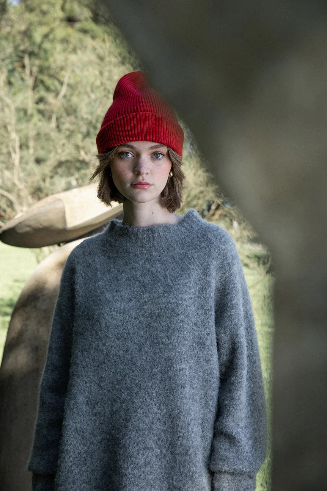 A person wearing the Hinoki Beanie – Poppy and a loose gray sweater stands outdoors near abstract stone sculptures, with trees in the background.