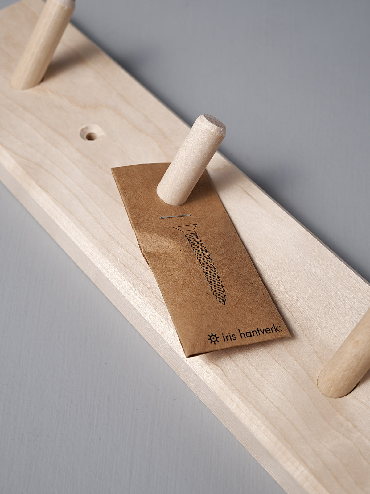 The Birchwood Peg Rail – 4 Hook by Iris Hantverk is crafted from untreated birch wood and showcases a Shaker-inspired aesthetic. It features four pegs and comes with a brown paper packet displaying a screw illustration alongside the words &quot;Iris Hantverk,&quot; epitomizing simple yet stylish craftsmanship.