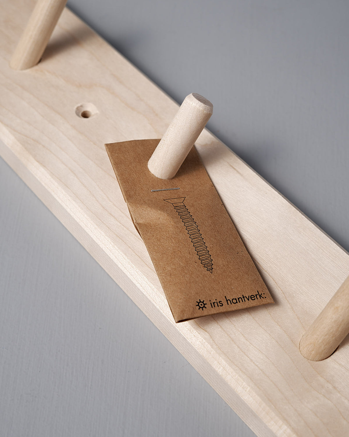 The Birchwood Peg Rail – 4 Hook by Iris Hantverk is crafted from untreated birch wood and showcases a Shaker-inspired aesthetic. It features four pegs and comes with a brown paper packet displaying a screw illustration alongside the words "Iris Hantverk," epitomizing simple yet stylish craftsmanship.