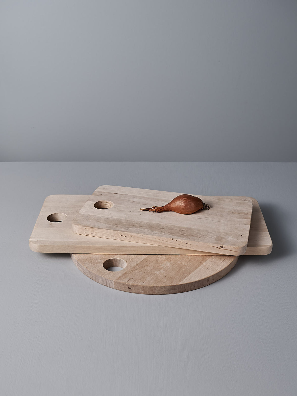 Three Birch Chopping Boards – Large by Iris Hantverk, ideal for use as a cheese board or serving tray, are stacked on a gray surface with a single shallot placed on top.