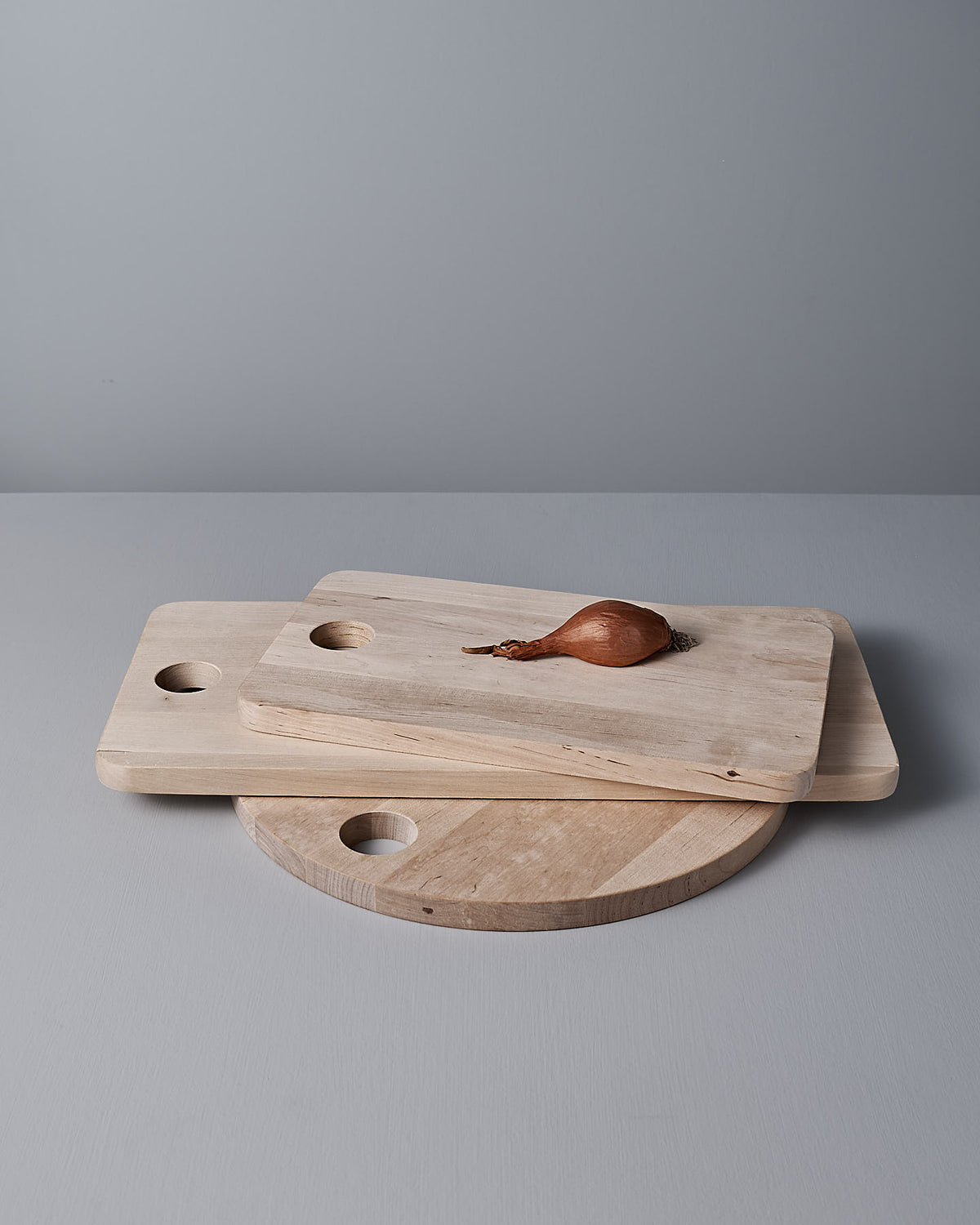 Three Birch Chopping Boards – Large by Iris Hantverk, ideal for use as a cheese board or serving tray, are stacked on a gray surface with a single shallot placed on top.