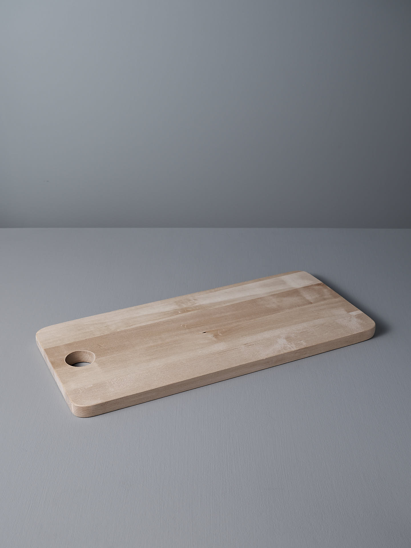 The Birch Chopping Board – Large by Iris Hantverk, with its rectangular shape, rounded edges, and small hole in one corner, lies flat on a gray surface against a matching background, doubling perfectly as a serving tray.