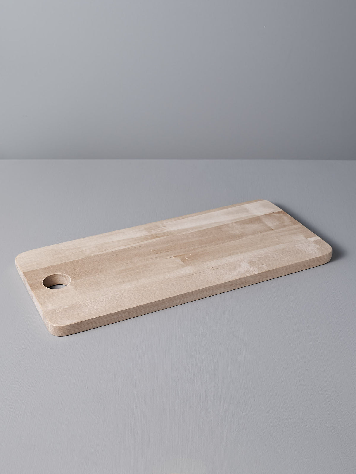 The Birch Chopping Board – Large by Iris Hantverk, featuring a rectangular design with rounded edges and a circular hole on one side, serves beautifully as a serving tray on the light gray surface.