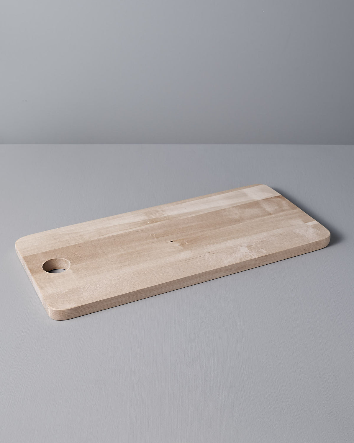 The Birch Chopping Board – Large by Iris Hantverk, featuring a rectangular design with rounded edges and a circular hole on one side, serves beautifully as a serving tray on the light gray surface.