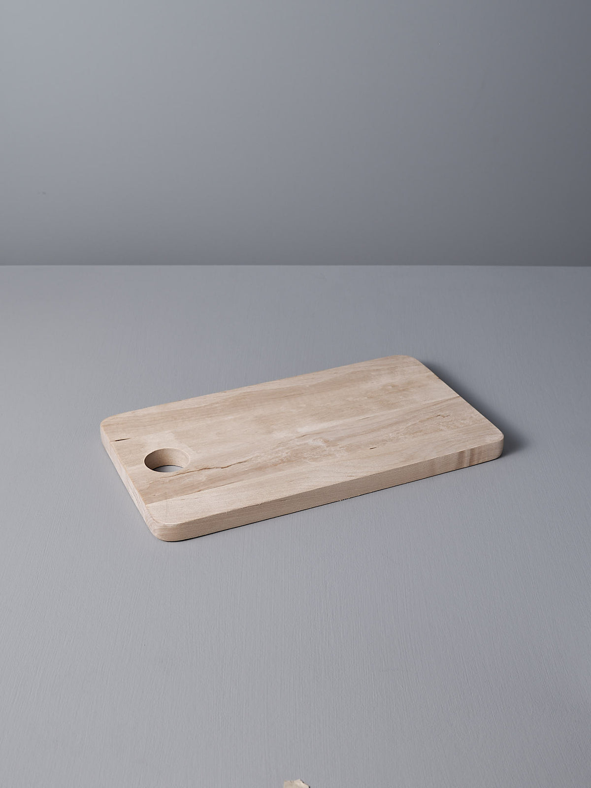 The Birch Chopping Board – Medium by Iris Hantverk is elegantly placed on a gray surface, featuring a circular hole as part of its design.