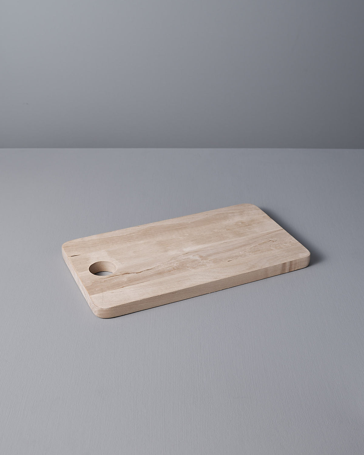 The Birch Chopping Board – Medium by Iris Hantverk is elegantly placed on a gray surface, featuring a circular hole as part of its design.