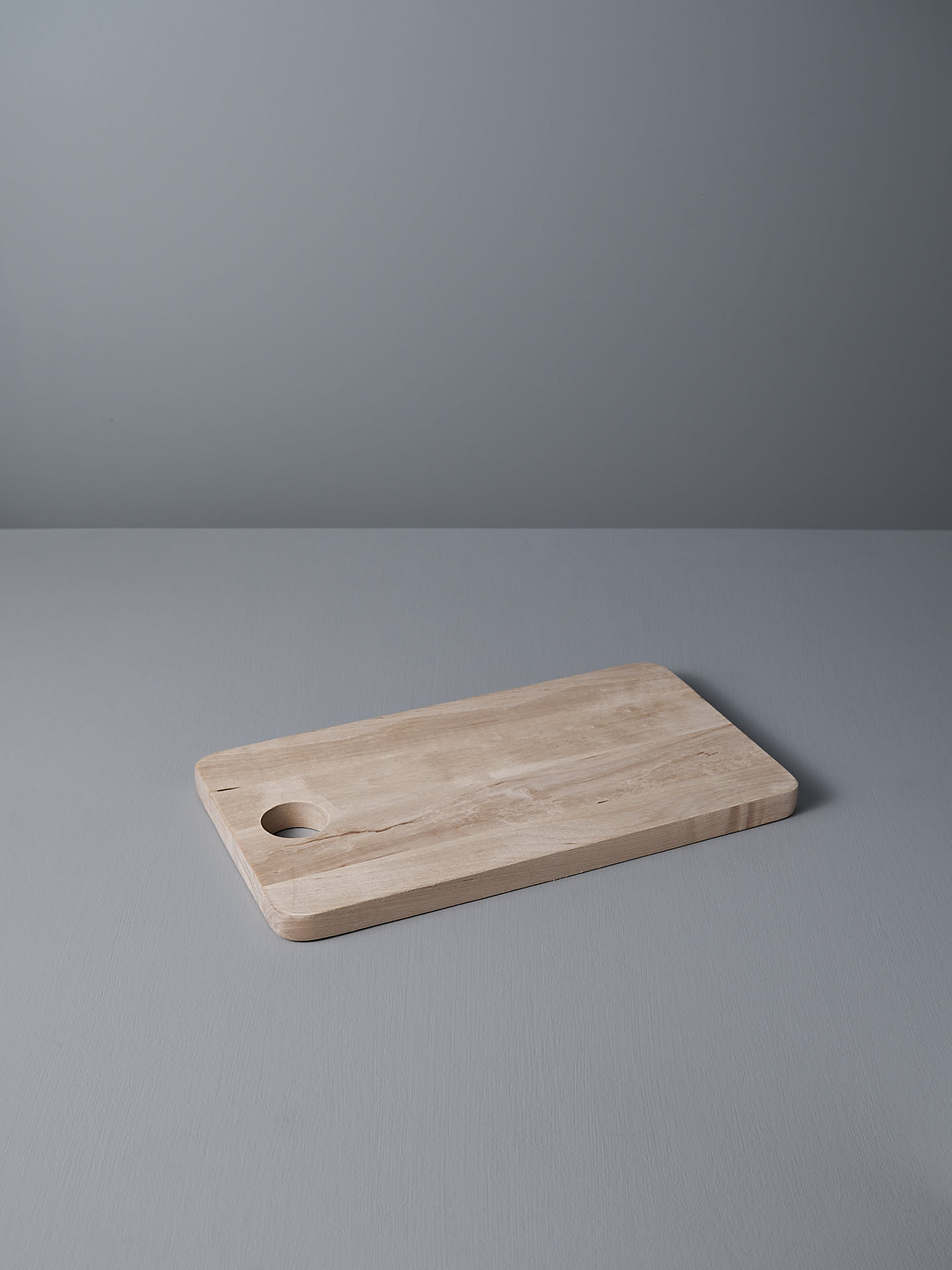The Birch Chopping Board – Medium from Iris Hantverk, a rectangular wooden board with rounded edges and a circular hole in one corner, ideal as a cheese board, is placed on a gray surface with a matching gray background.
