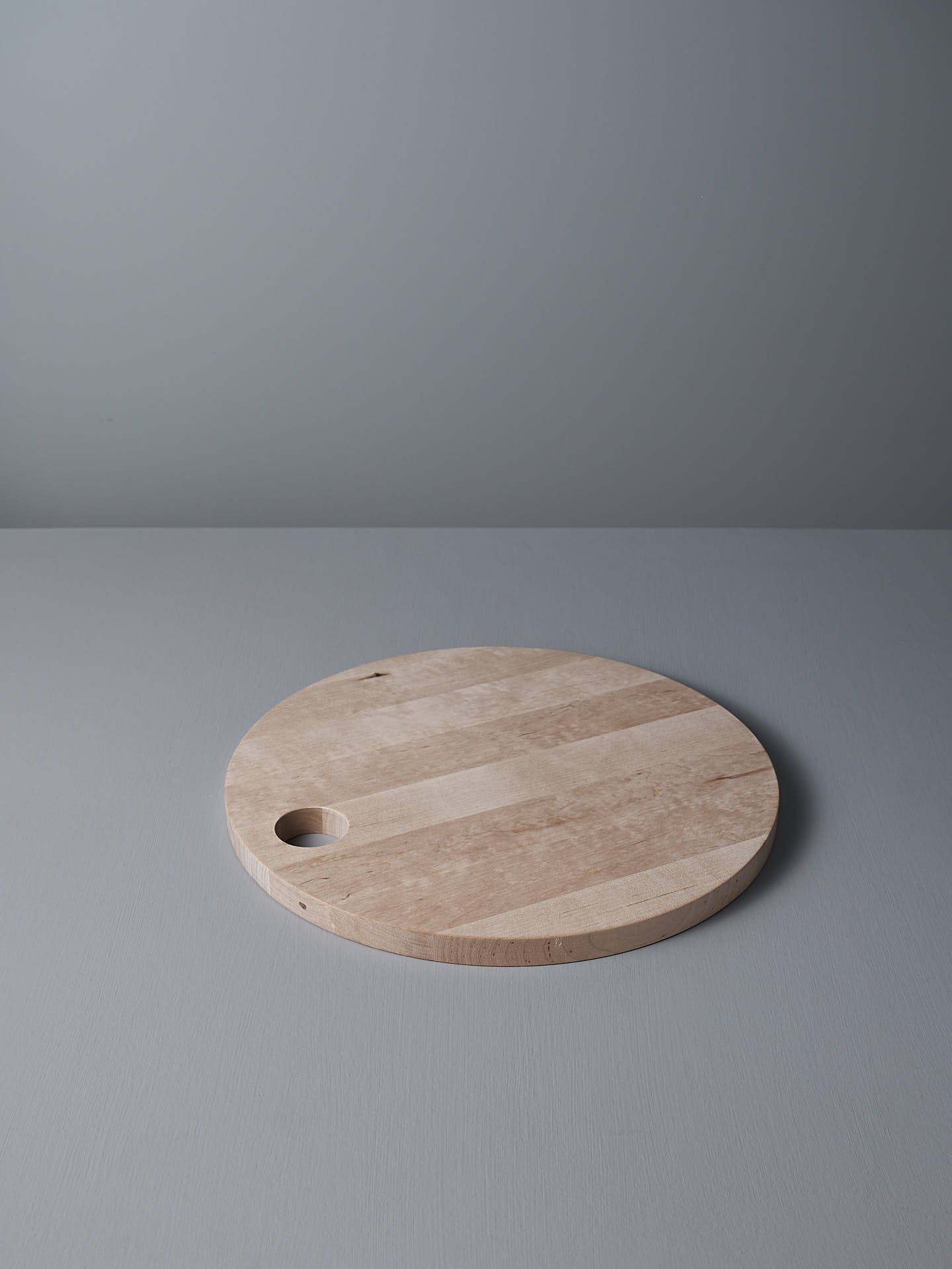 The Iris Hantverk Birch Chopping Board – Round, treated with food-grade oil for durability and featuring a small hole near the edge, rests on a gray surface against a matching gray background.