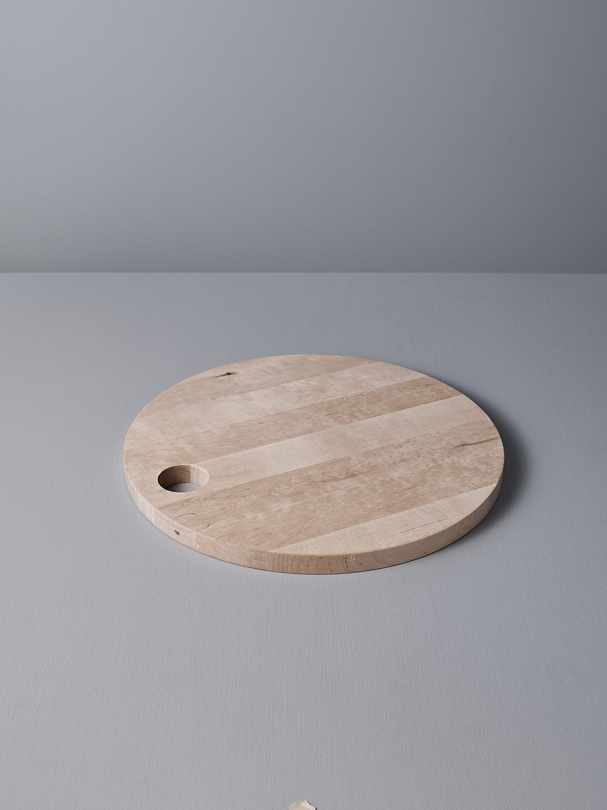 The Iris Hantverk Birch Chopping Board – Round, made from untreated birch, features a hole near the edge and rests on a light gray surface.