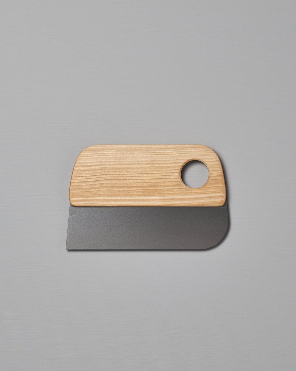 An Iris Hantverk Dough Scraper with a birch wooden handle and a circular hole, elegantly placed on a gray surface.