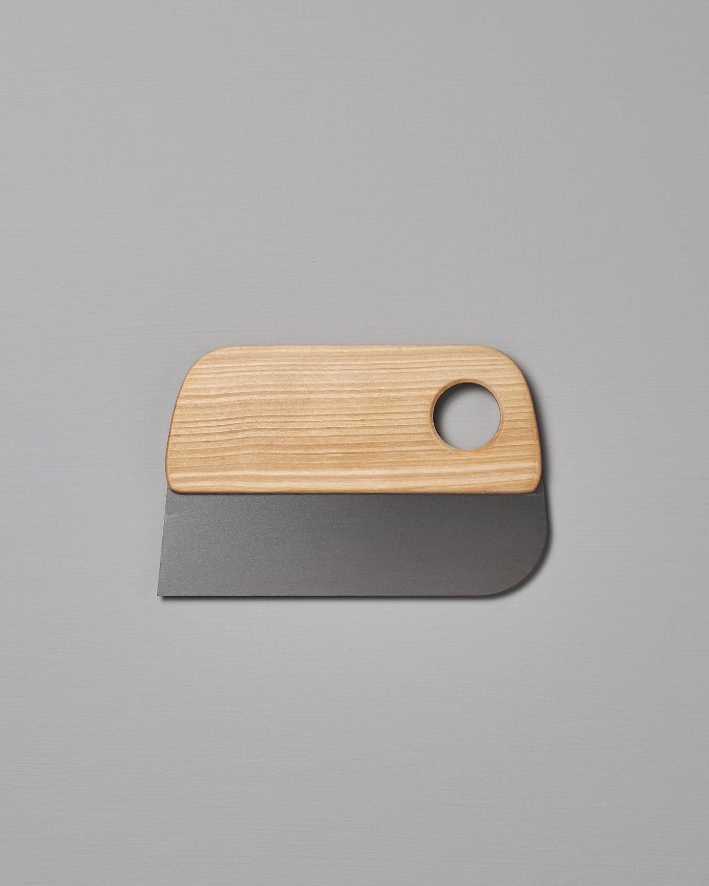 An Iris Hantverk Dough Scraper with a stainless steel blade and a circular hole in the handle, placed on a plain gray surface. This essential baking tool is perfect for all your pastry needs.