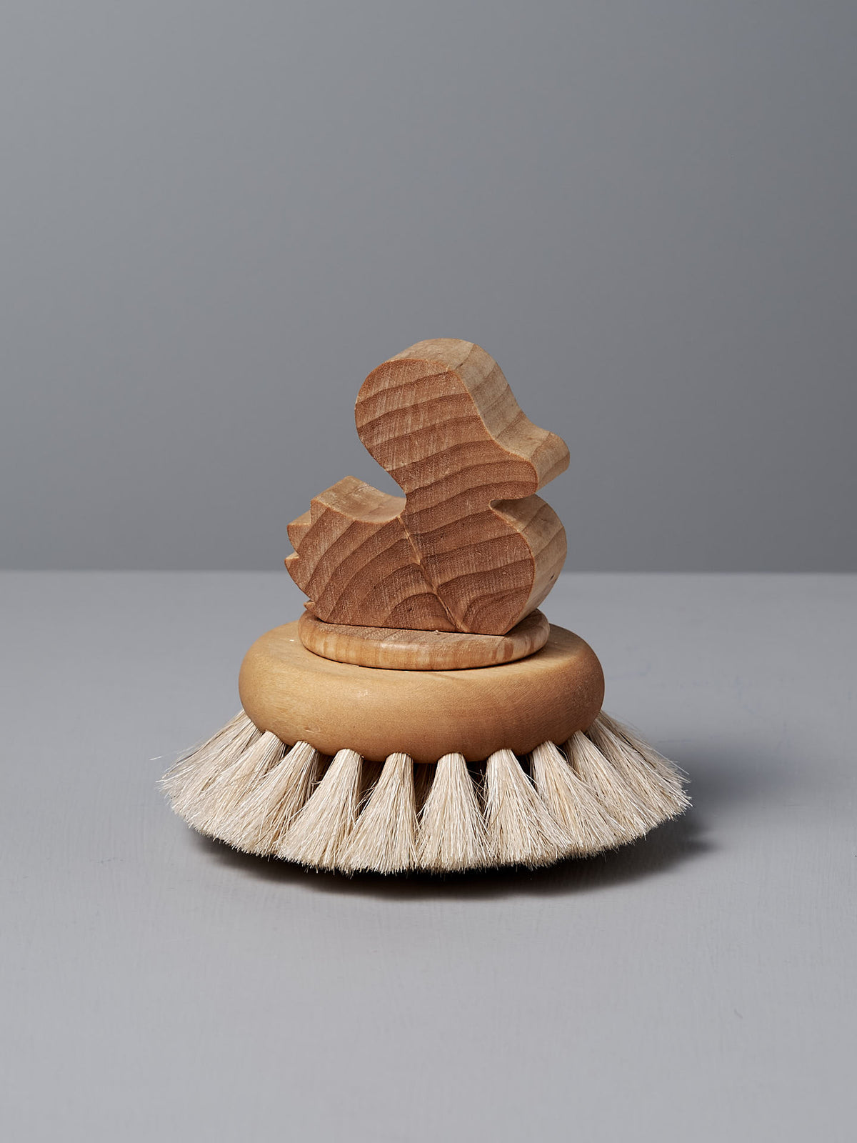 The Duck Bath Brush – Soft by Iris Hantverk features a wooden duck-shaped handle atop a circular scrub brush with gentle bristles, expertly crafted and displayed on a gray surface.