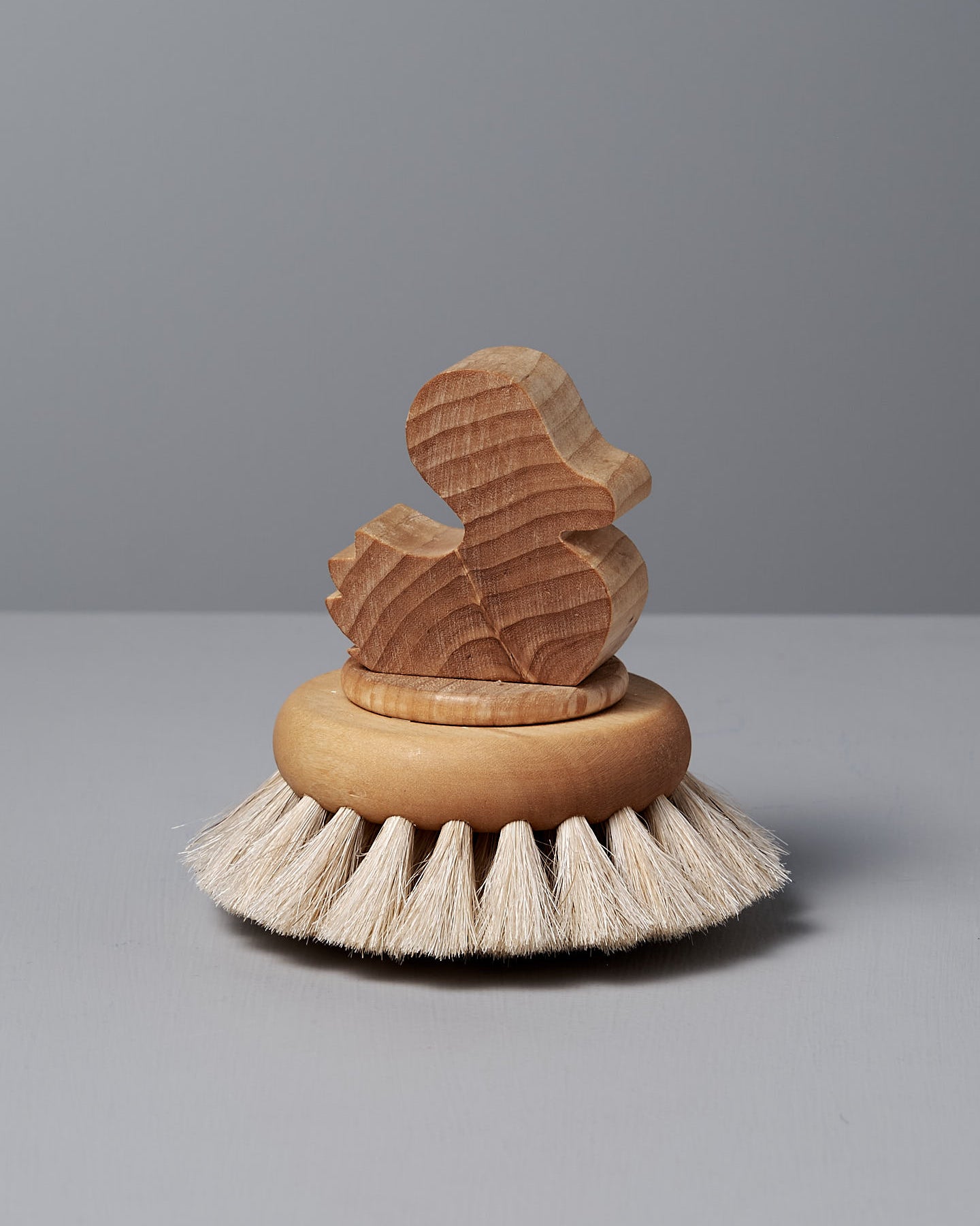 The Duck Bath Brush – Soft by Iris Hantverk features a wooden duck-shaped handle atop a circular scrub brush with gentle bristles, expertly crafted and displayed on a gray surface.