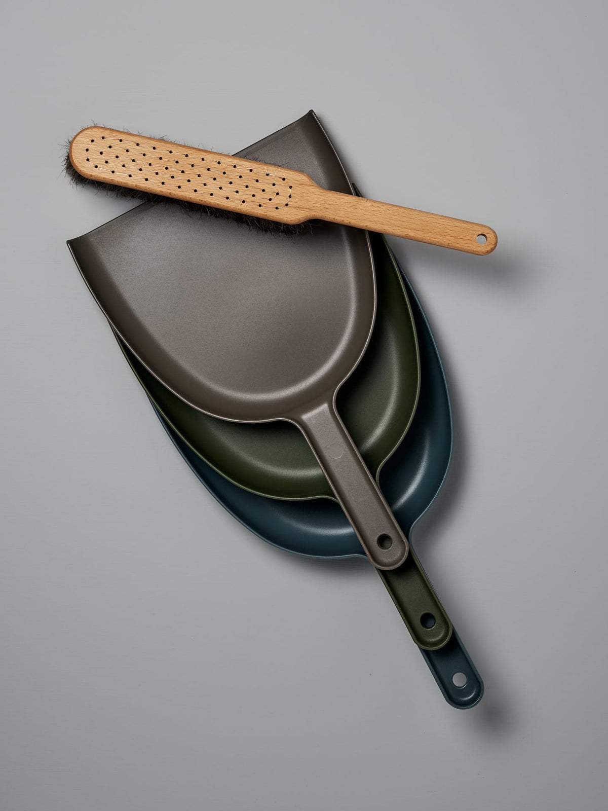 A set of three nested dustpans in shades of gray, green, and blue features an oiled beech brush with horsehair bristles resting on top, against a sleek gray background. The Dustpan &amp; Brush Set – Umber by Iris Hantverk is a stylish and functional addition to any cleaning routine.