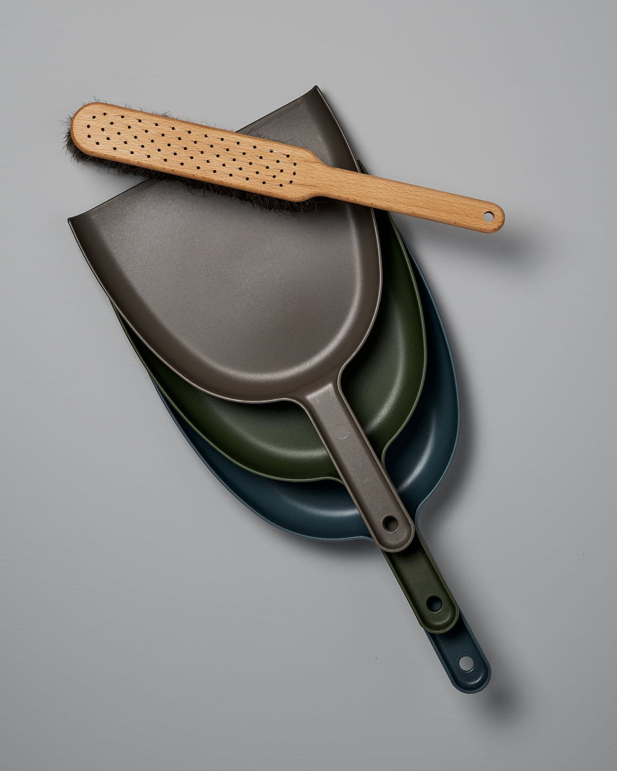 A set of three nested dustpans in shades of gray, green, and blue features an oiled beech brush with horsehair bristles resting on top, against a sleek gray background. The Dustpan & Brush Set – Umber by Iris Hantverk is a stylish and functional addition to any cleaning routine.