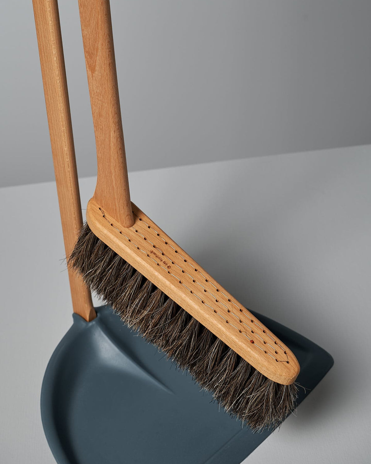 Wooden-handled broom with bristles resting on a matching blue dustpan, both part of an eco-friendly cleaning tools set positioned on a smooth, light grey surface. The Long Handled Dustpan & Brush Set – Petrol Blue from Iris Hantverk adds a touch of elegance to your cleaning routine.