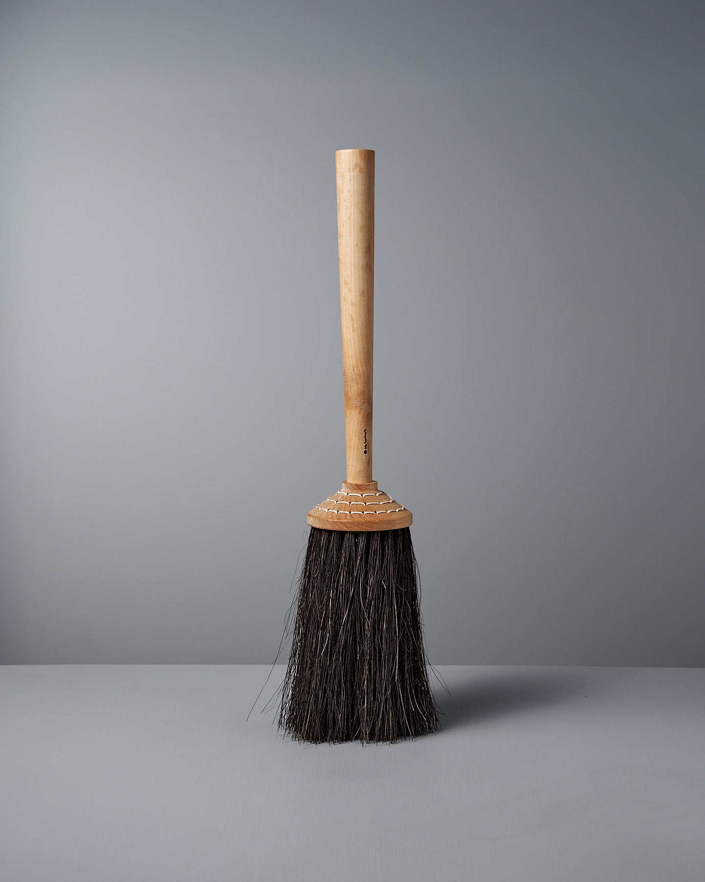 The Iris Hantverk Short Handle Porch Broom, featuring a wooden handle made of birch and dark Arenga bristles, stands upright against a plain grey background.
