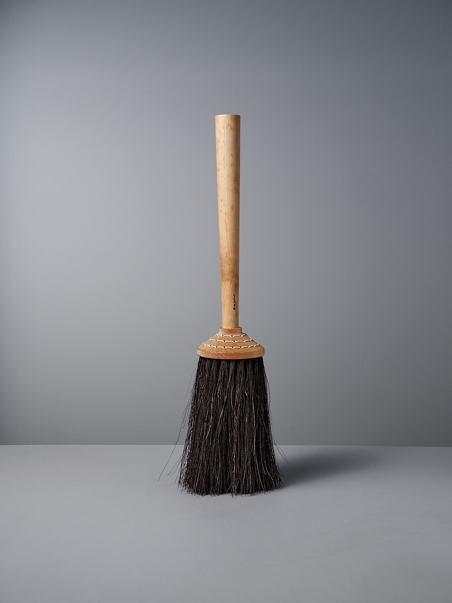 The Iris Hantverk Short Handle Porch Broom, featuring a wooden handle made of birch and dark Arenga bristles, stands upright against a plain grey background.