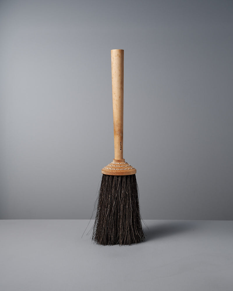 The Iris Hantverk Short Handle Porch Broom, featuring a wooden handle made of birch and dark Arenga bristles, stands upright against a plain grey background.