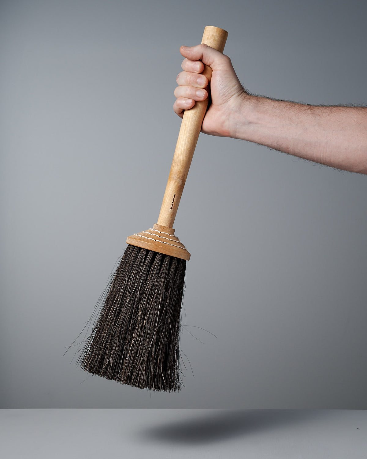 A hand holding the Short Handle Porch Broom – Birch & Arenga, featuring dark Arenga bristles by Iris Hantverk, against a gray background.