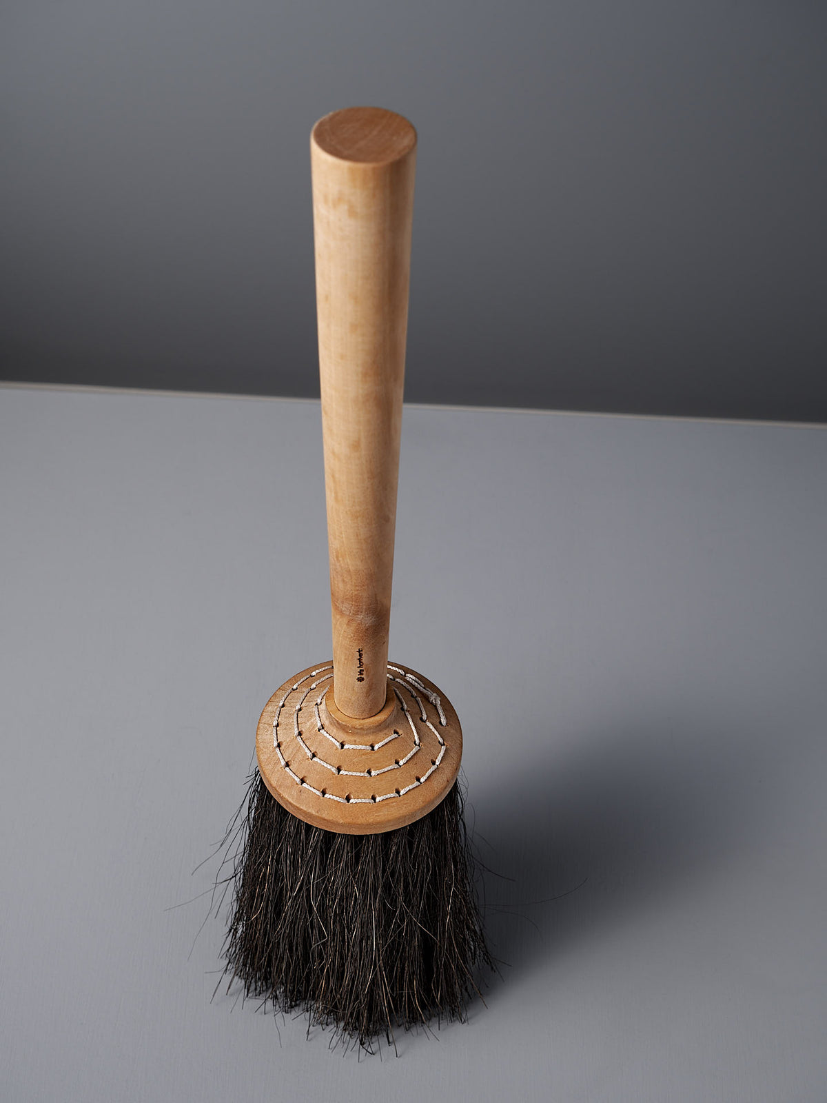 An Iris Hantverk Short Handle Porch Broom, made with birch wood and Arenga bristles, stands elegantly upright on a gray surface.