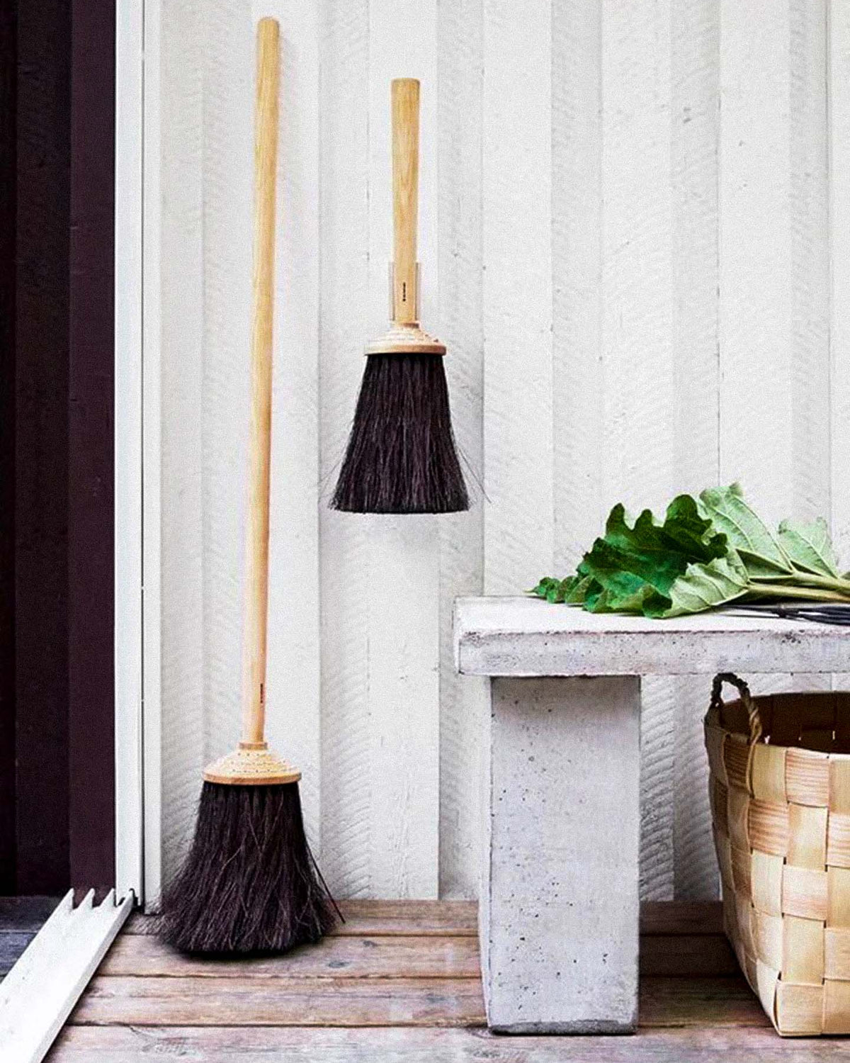 Two Iris Hantverk Short Handle Porch Brooms, crafted with birch and arenga, lean against a white wall. One broom rests on the floor showcasing its durable arenga bristles, while the other hangs neatly on the wall. A concrete bench adorned with green leaves and accompanied by a woven basket completes the serene setup.