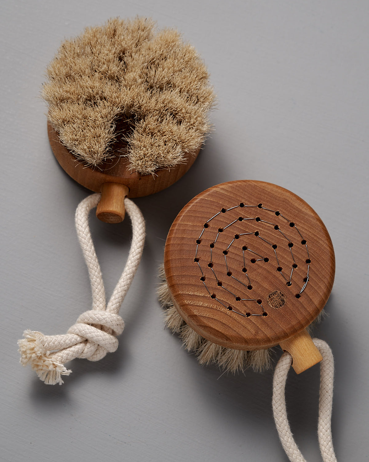 Two Iris Hantverk Puck Bath Brushes, featuring natural horsehair bristles and wooden handles with rope loops, are elegantly displayed on a gray surface, highlighting their artisanal craftsmanship.