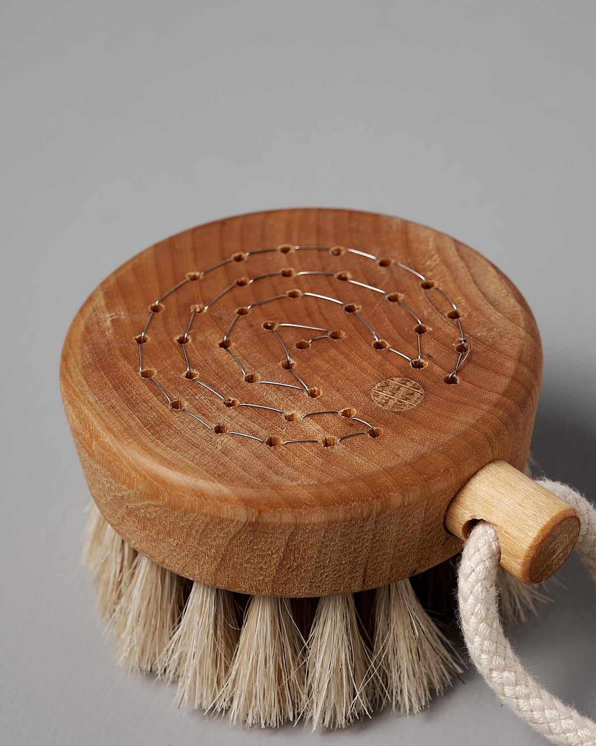 The Puck Bath Brush by Iris Hantverk is a round wooden bath brush with natural horsehair bristles and a string loop handle, featuring a decorative pattern made from small nails and string on the top surface.