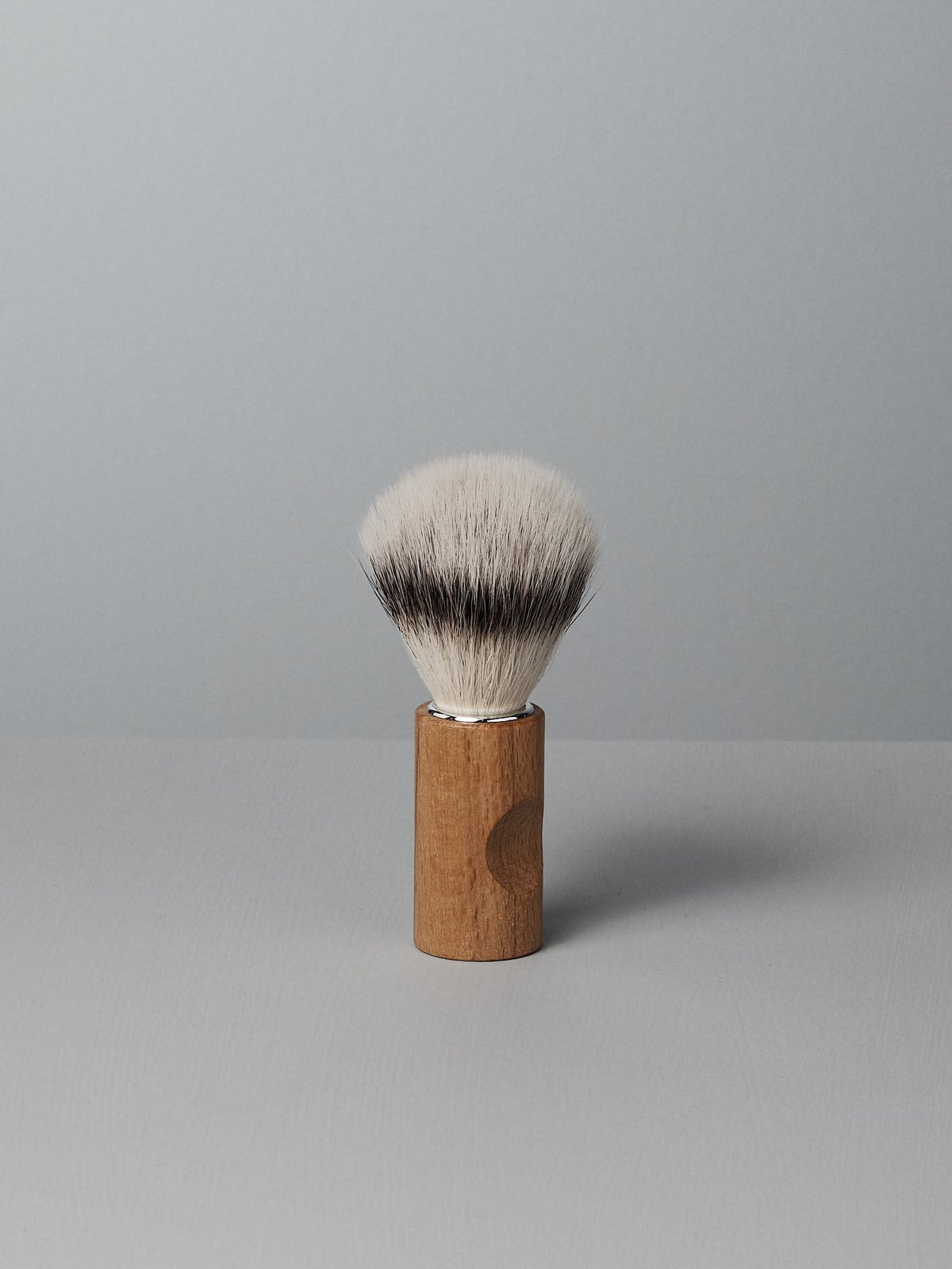 A Shaving Brush – Silver Tip by Iris Hantverk with a wooden handle and cruelty-free synthetic fibres stands upright on a light gray surface against a light gray background.