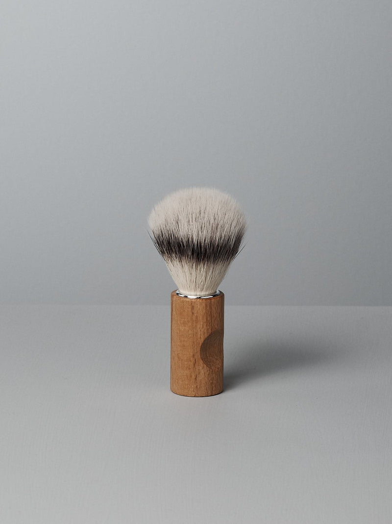 A Shaving Brush – Silver Tip by Iris Hantverk with a wooden handle and cruelty-free synthetic fibres stands upright on a light gray surface against a light gray background.