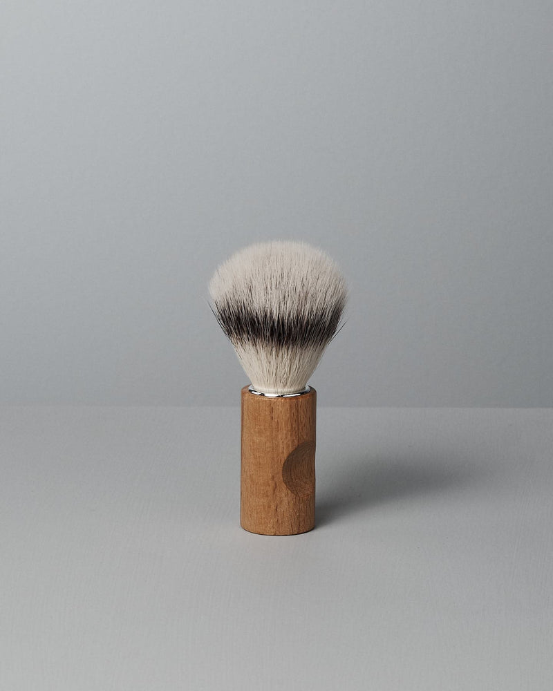 A Shaving Brush – Silver Tip by Iris Hantverk with a wooden handle and cruelty-free synthetic fibres stands upright on a light gray surface against a light gray background.