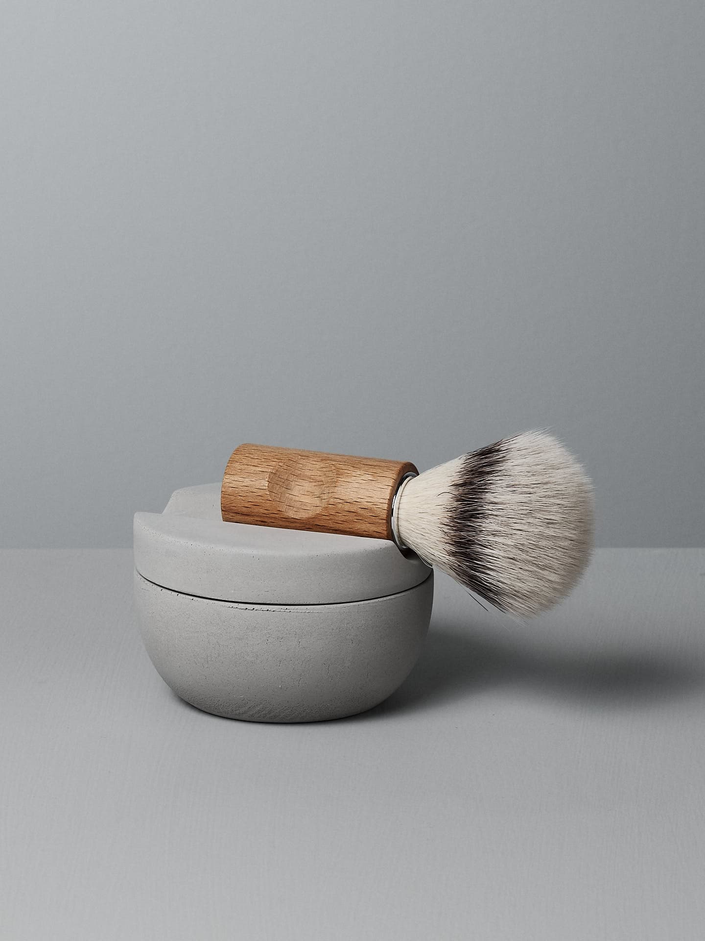 An Iris Hantverk Shaving Brush & Cup Set, with Cedarwood Soap rests atop a closed grey shaving cup against a grey background.