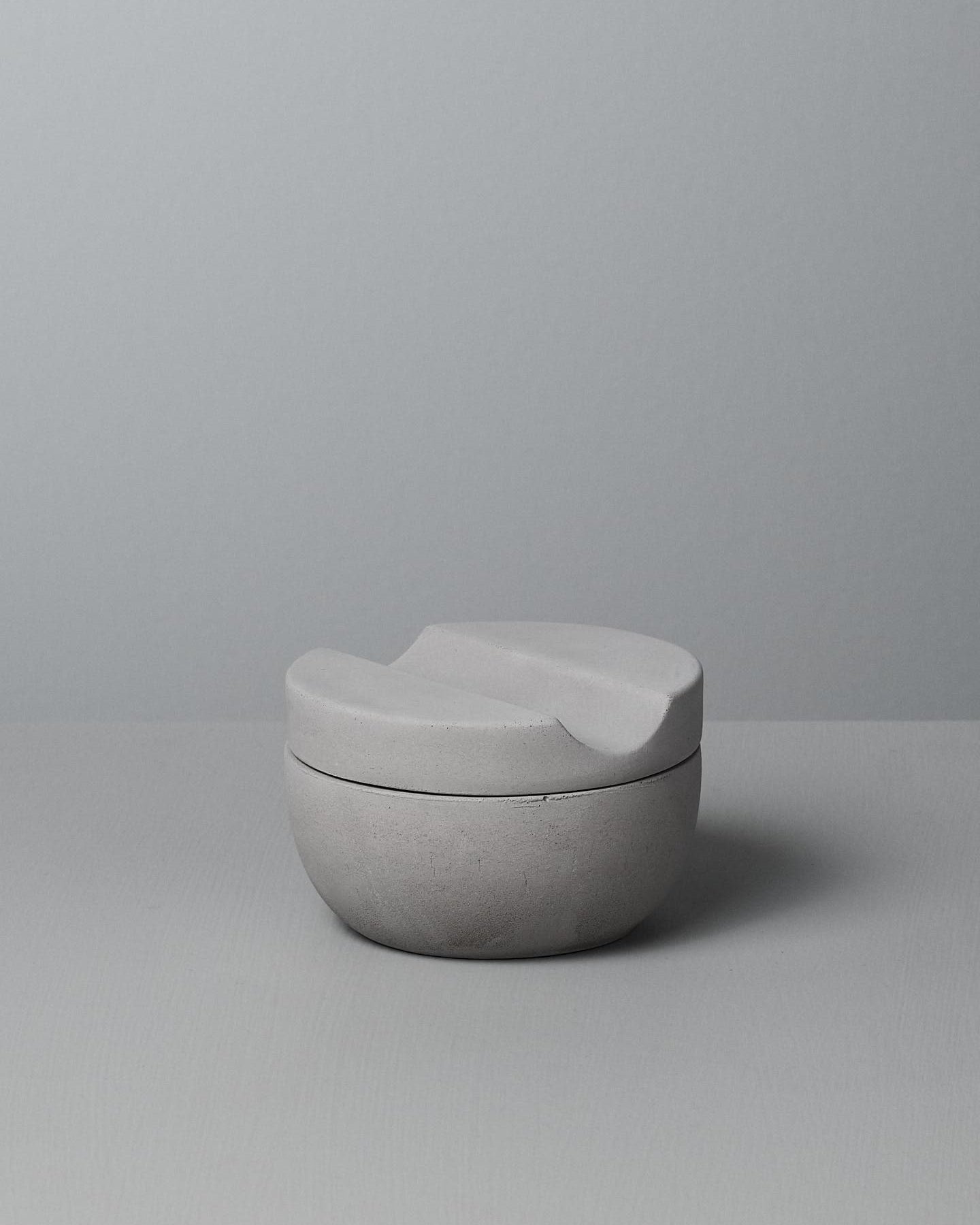 An Iris Hantverk Shaving Cup with Cedarwood Shaving Soap, a minimalist, grey ceramic cup with a curved lid, rests on a soft concrete surface, perfect for holding your cedarwood shaving soap.